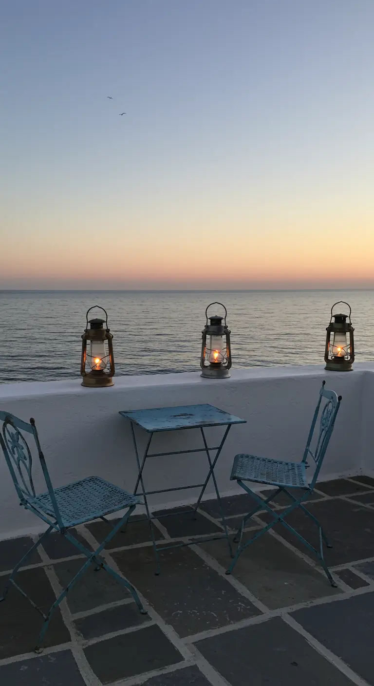 Balcón minimalista frente al mar con sillas de hierro azul y tres faroles sobre un murete al atardecer