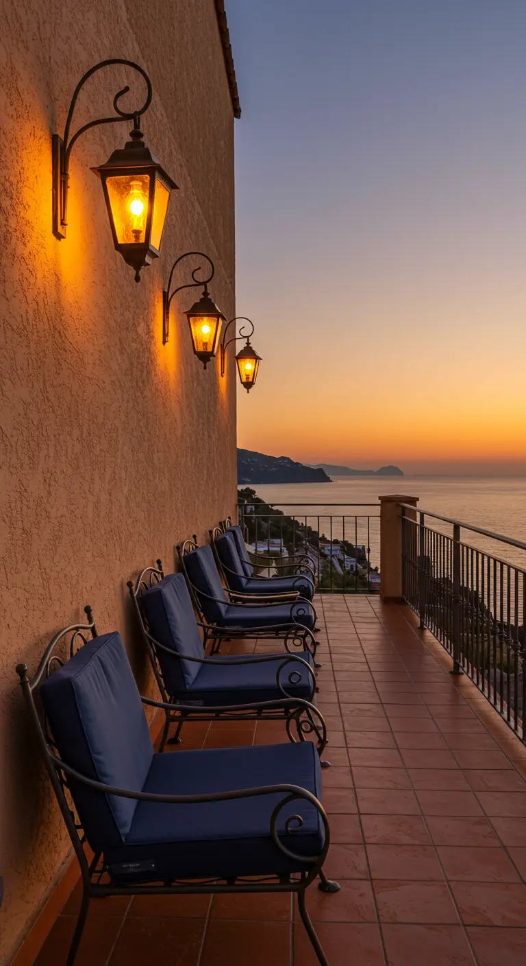 Terraza con vistas al mar, sillas de hierro en fila y faroles de pared encendidos al atardecer