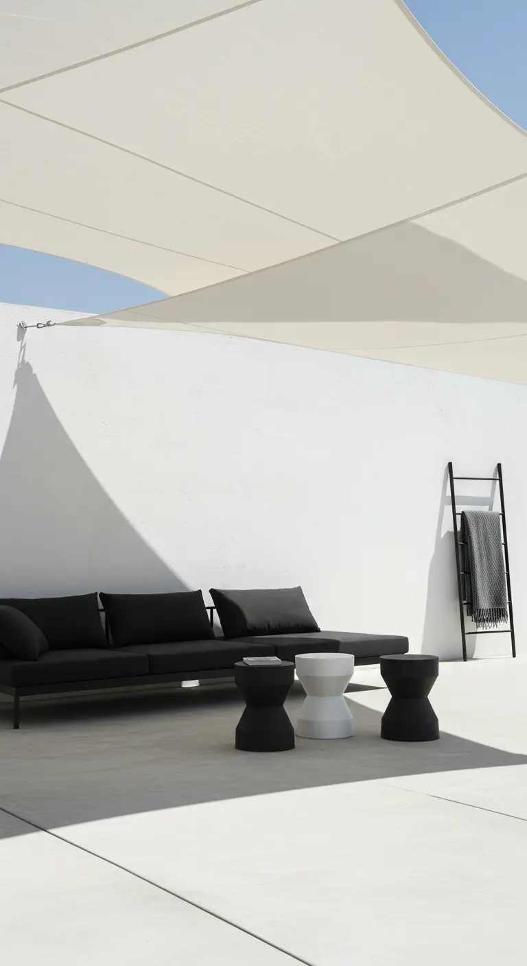Patio minimalista con paredes blancas, sofá negro, mesas auxiliares y una vela de sombra blanca.