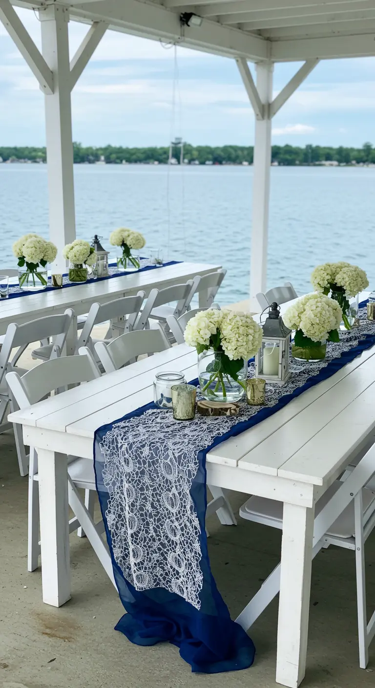 Mesas de boda blancas con un camino de encaje azul marino y flores blancas.