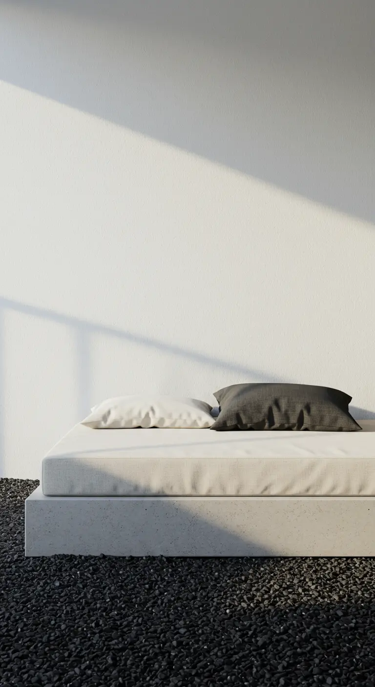 Una cama de día baja y blanca con cojines sobre un suelo de grava negra en un espacio minimalista.