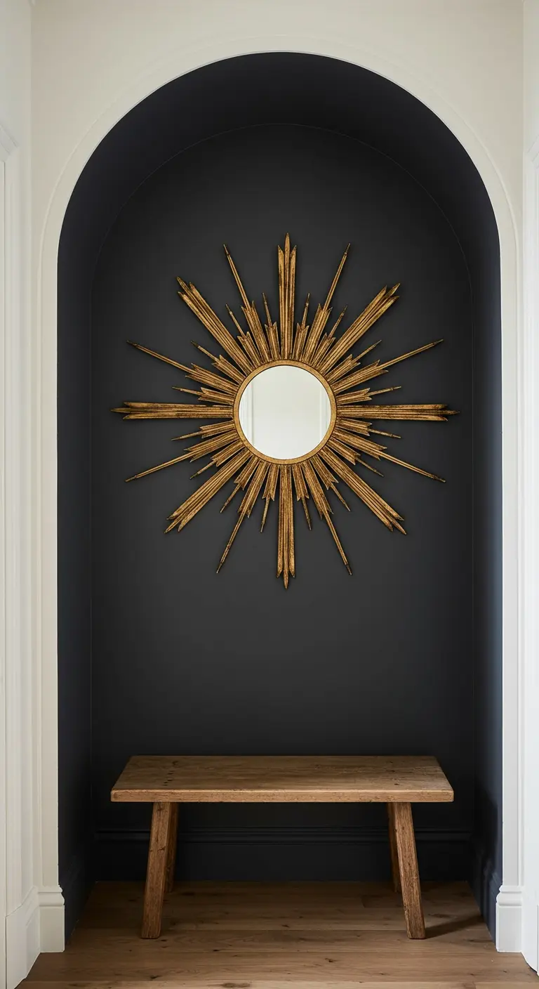 Nicho de pared pintado de oscuro con un espejo de sol dorado y un banco de madera debajo.
