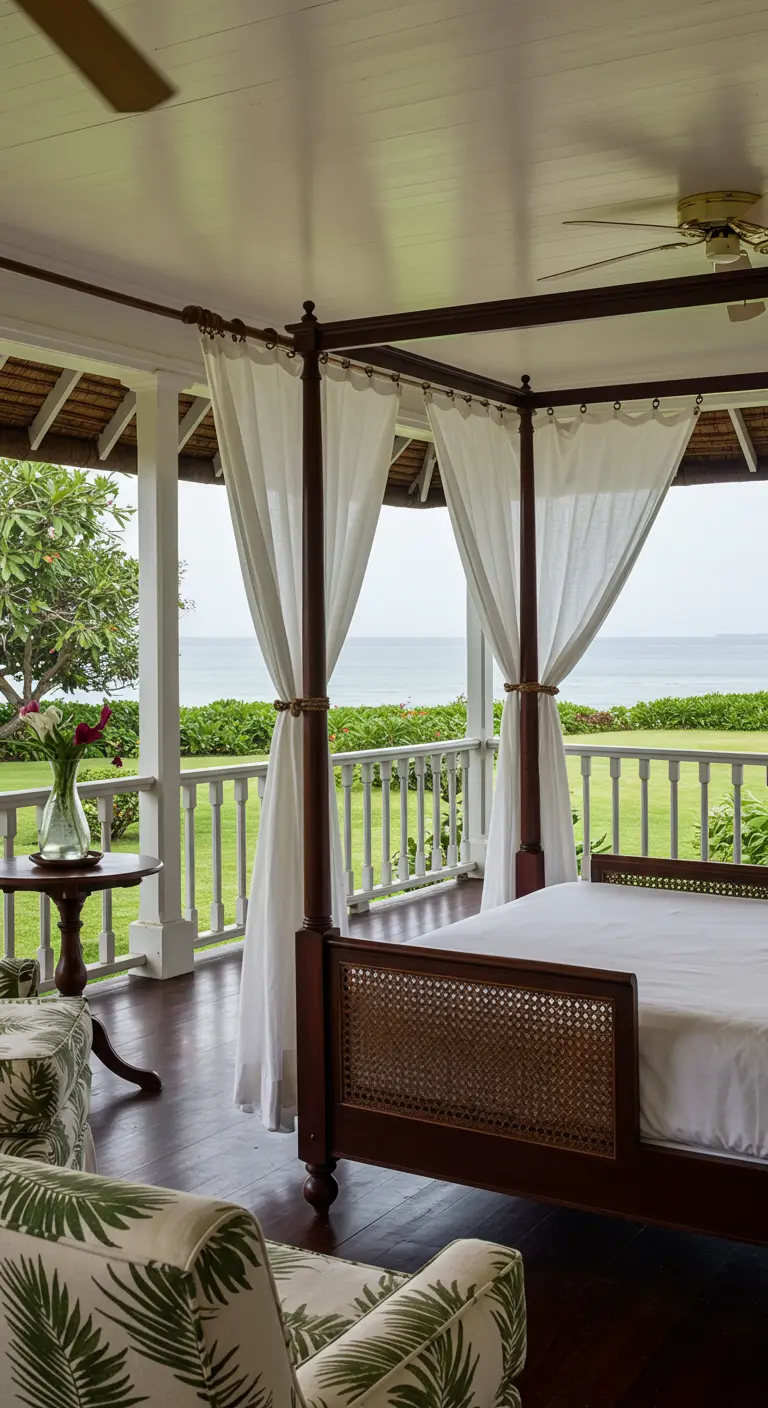 Cama con dosel de madera oscura y cortinas blancas en un porche con vistas a un jardín y al mar.