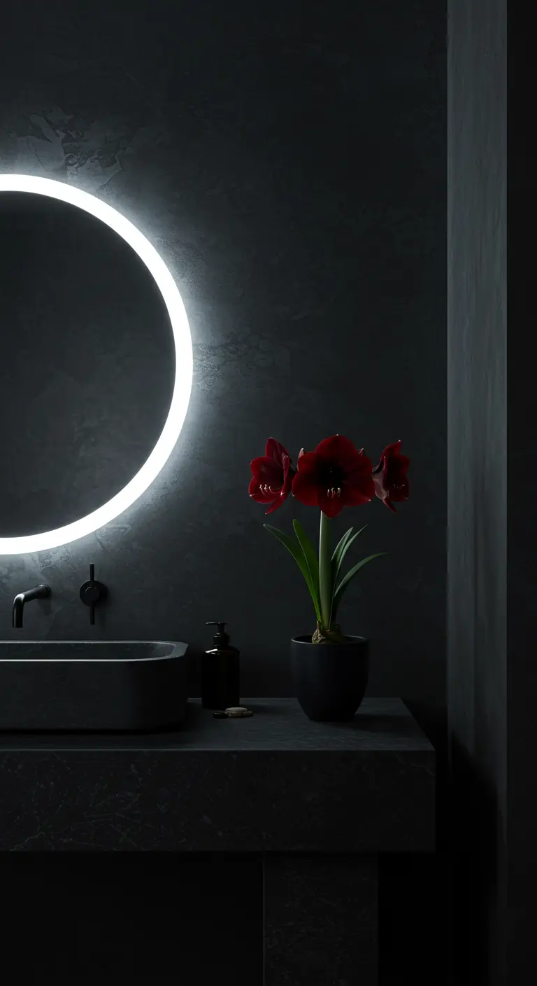 Baño oscuro y minimalista con un espejo redondo iluminado y una planta de amarilis roja.