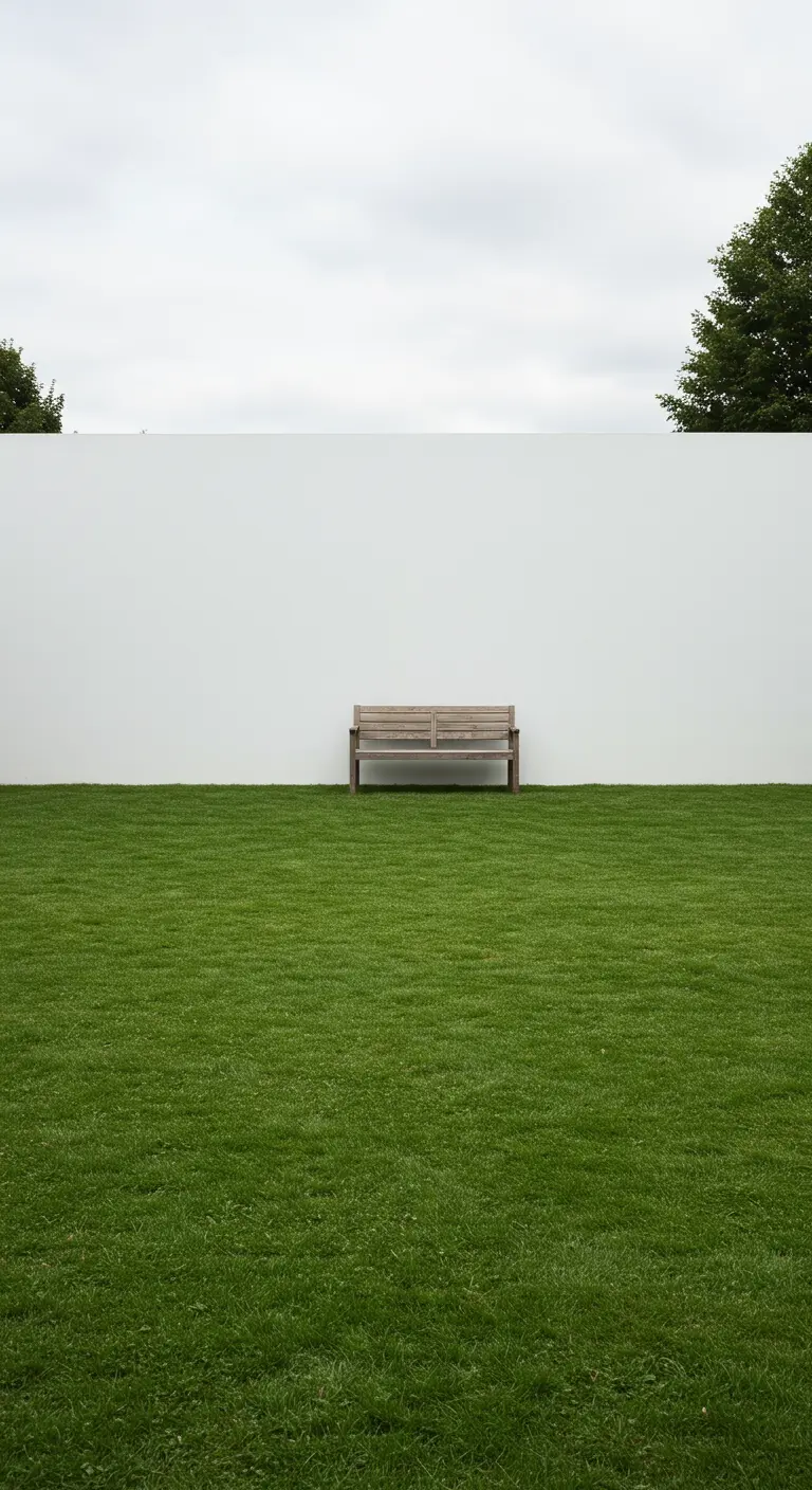 Un único banco de madera colocado sobre un césped verde inmaculado frente a un alto muro blanco.