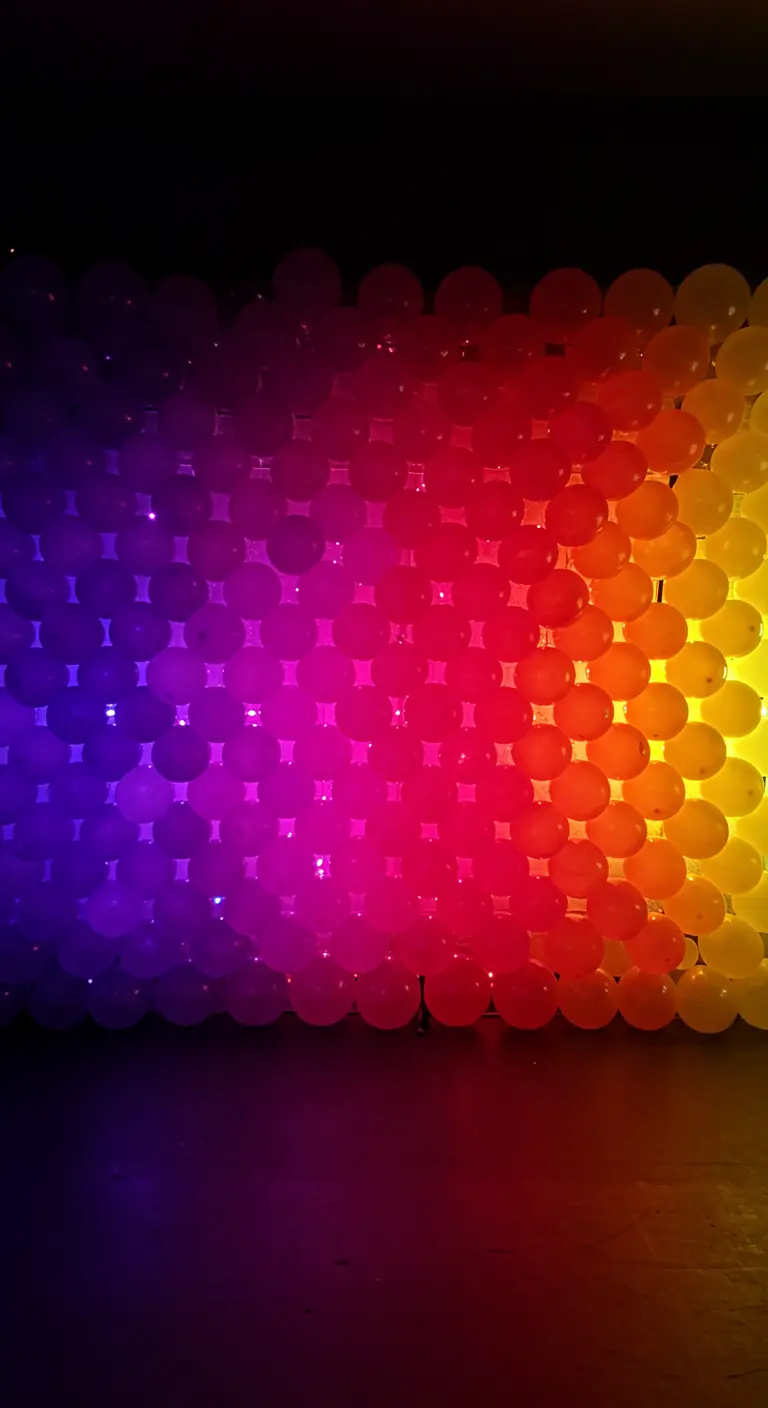 Pared hecha de globos de colores del arcoíris, iluminada desde atrás en un degradado.