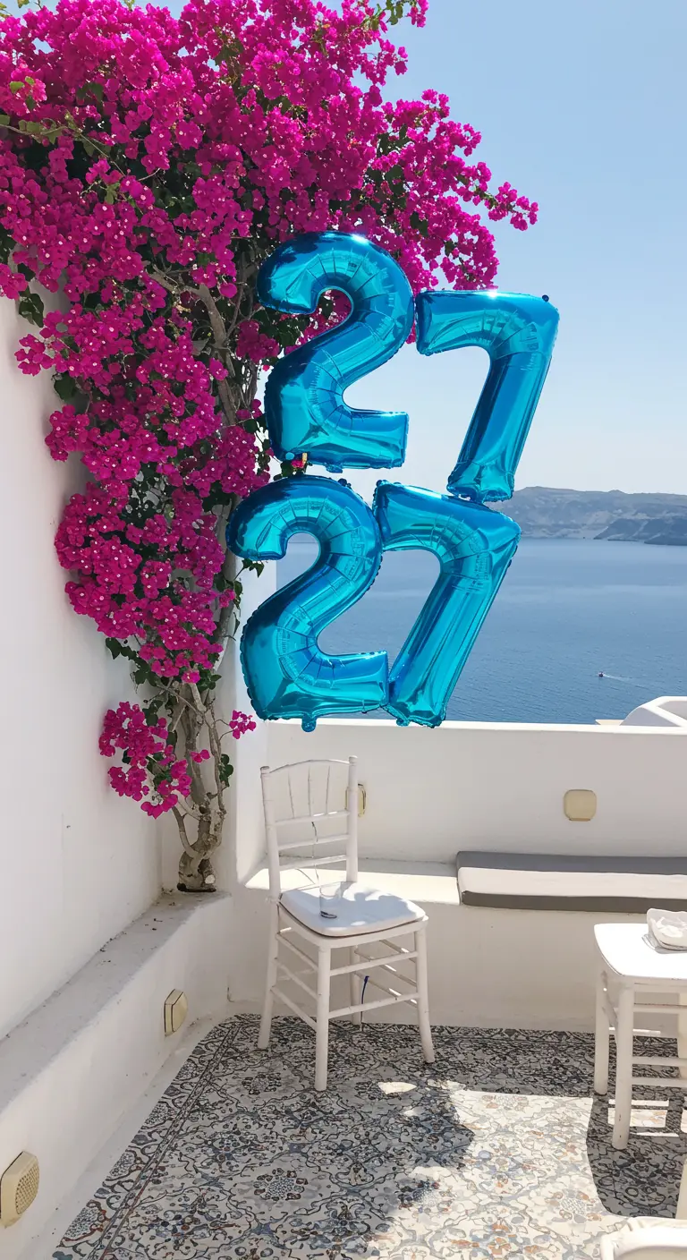 Globos azules del número 27 en un balcón blanco con una buganvilla fucsia y vistas al mar.