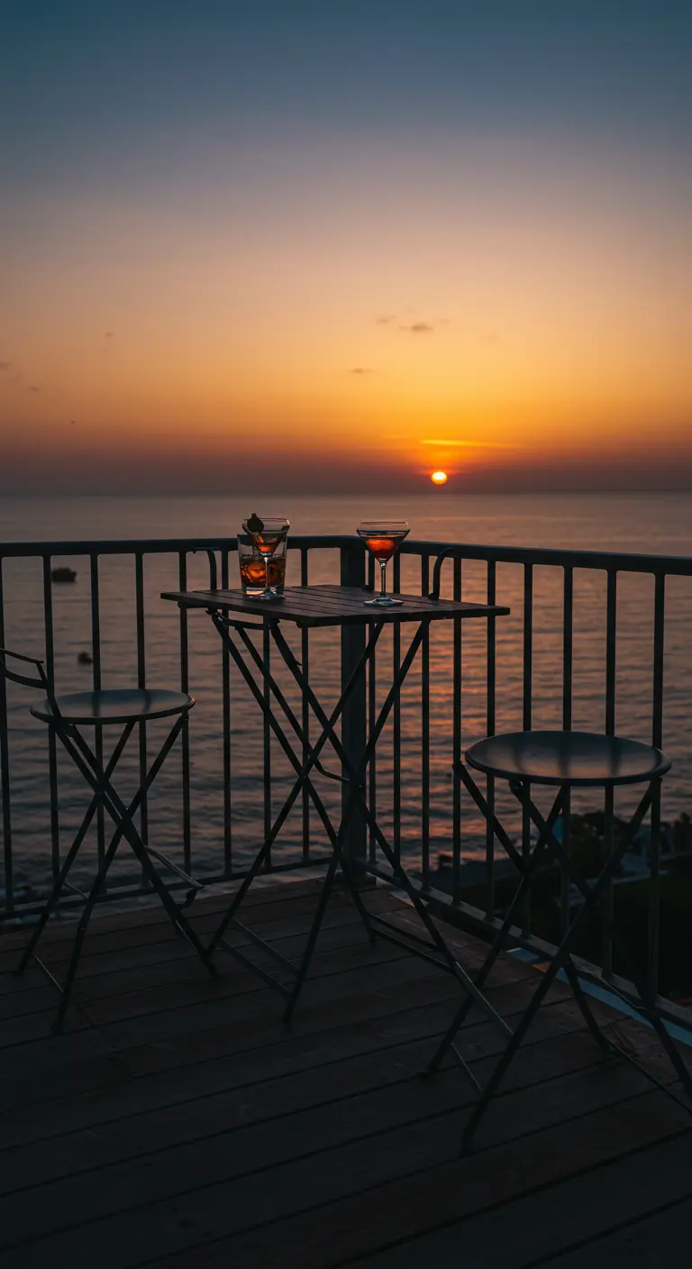 Silueta de una mesa alta y dos taburetes plegables en un balcón frente a una puesta de sol en el mar.
